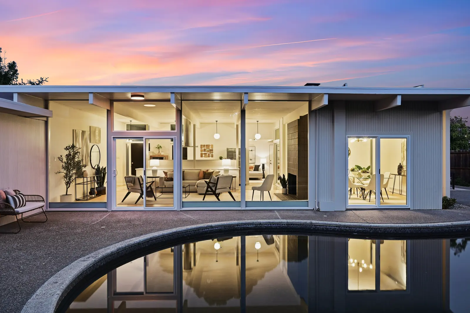 A mid-century home at dusk, its glass walls lit from inside and mirrored in a curved pool